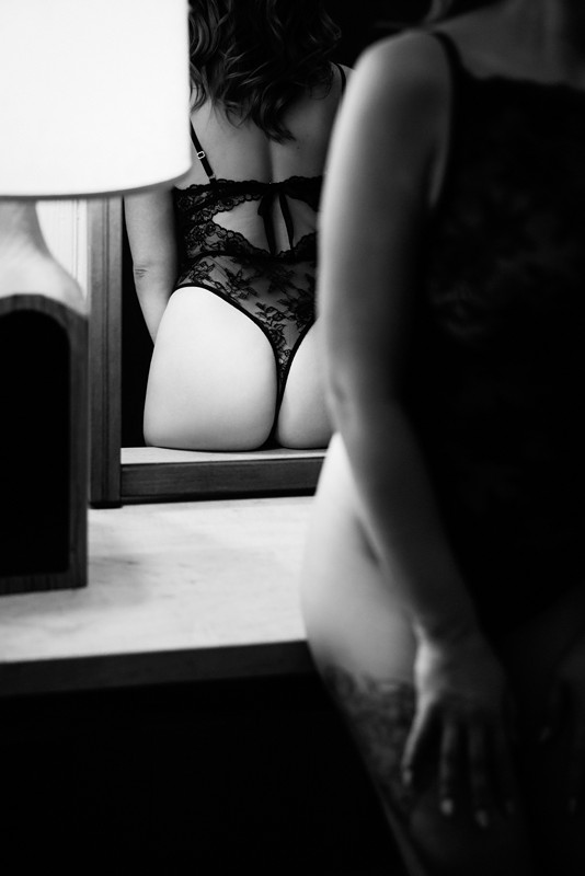 a woman poses on a desk in a black thong bodysuit reflected in a mirror behind her during her boudoir photoshoot at a chicago boudoir studio